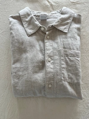 Beige linneskjorta med bröstficka - Stilren beige skjorta i linne med klassisk krage och bröstficka som kommer passa perfekt till sommaren. Skjortan har långa ärmar och knäppning framtill med vita knappar. Perfekt för en avslappnad men snygg look i värmen.