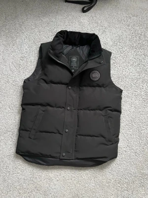 Svart dunväst från Canada Goose - Snygg svart dunväst från Canada Goose med hög krage och diskret logomärke på bröstet. Västen har två sidofickor och stängs med både dragkedja och tryckknappar. Perfekt för lager-på-lager och håller dig varm under kyliga dagar.