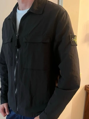 Svart overshirt från Stone Island - Snygg svart overshirt från Stone Island med dragkedja framtill och två bröstfickor med lock. Klassisk patch med logga på vänster ärm. Overshirten har en rak passform och är tillverkad i ett lätt material som passar perfekt till lager-på-lager.