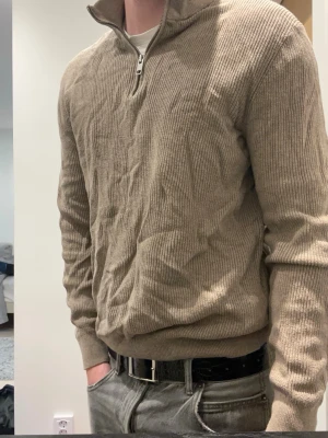 Beige stickad halvzip från H&M - Säljer en beige stickad tröja från H&M med halvzip och hög krage. Tröjan har ribbstickad struktur och långa ärmar. Perfekt för lager på lager och enkel att matcha med olika outfits.