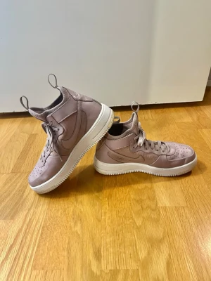 Nike Air Force 1 - Storlek 40 - Ett par Nike Air Force 1 sneakers i en tilltalande ljuslila färg. Skorna har en klassisk design med snörning och en bekväm passform. De är utrustade med en robust sula som ger bra grepp och stöd. Perfekta för vardagsbruk. Skorna är i bra skick däremot är sulan lite mer sliten jämfört med andra delar av skon som ni ser på bilderna.