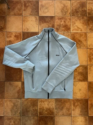 Ljusgrå zip hoodie från BOSS - Snygg ljusgrå hoodie från BOSS med hel dragkedja framtill och diskret BOSS-logga på bröstet. Hoodien har hög krage, långa ärmar och två fickor med dragkedja. Tillverkad i mjukt material för en bekväm känsla. Perfekt för en avslappnad och stilren look.