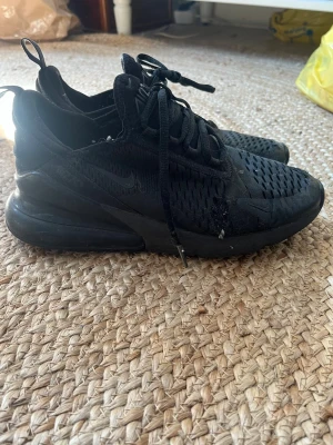Svarta Nike Air Max 270 sneakers - Säljer ett par helsvarta Nike Air Max 270 sneakers med mesh-ovandel och synlig Air-enhet i hälen. Skorna har klassisk Nike-logga på sidan, snörning och dragflik bak. Perfekta för dig som gillar sportig och stilren look.