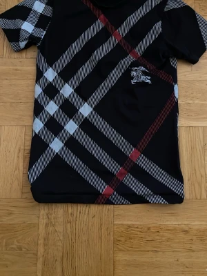 Burberry Svart rutig t-shirt - Snygg svart t-shirt med stort rutigt mönster i vitt och rött över hela plagget. T-shirten har korta ärmar och ett litet tryck på bröstet. Perfekt för dig som gillar streetstyle och vill sticka ut lite extra.