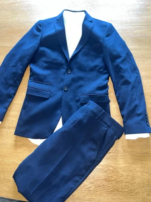 Marinblå kostym Selected Homme slim - Snygg marinblå kostym från Selected Homme i slim fit-modell. Kavajen har två knappar, klassiska fickor med lock och matchande byxor. Perfekt för dig som vill ha en stilren och modern look. Materialet är mjukt och följsamt, vilket ger en bekväm känsla. Använd en gång. Jacka - xs/s,   Skjorta - xs/36,  Byxor - 46. 