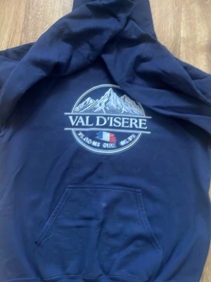 Mörkblå Val d'Isere hoodie - Mörkblå hoodie med tryck av berg och texten 'Val d'Isere' samt 'France Ski Club' på bröstet. Klassisk känguruficka framtill och stor huva. Tillverkad i mjukt bomullsmaterial, perfekt för chill dagar eller när du vill ha en sportig look.