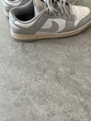 Nike Dunk Low gråvita sneakers - Nike Dunk Low sneakers i grått och vitt med klassisk swoosh på sidan. Ovandel i läder och mesh, låga med vadderad plös och snörning. Snygg retrostil som passar till streetwear och casual outfits.
