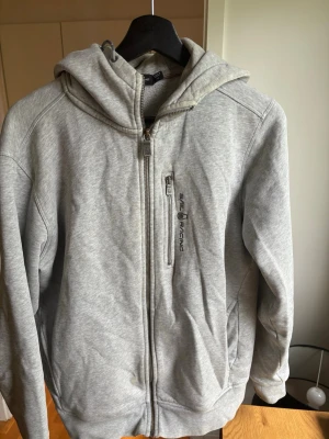 Grå hoodie från Sail Racing - Snygg grå hoodie från Sail Racing med dragkedja framtill och en bröstficka med logga. Mjuk insida i fleece och klassisk huva. Perfekt för en chill och sportig stil.