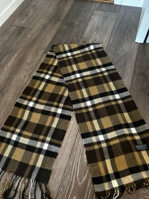 Rutig halsduk från Burberrys - Snygg rutig halsduk från Burberrys i brunt, beige, svart och vitt. Tillverkad i 100% lammull och har klassiskt rutmönster med fransar i ändarna. Perfekt accessoar för att lyfta din outfit med en tidlös brittisk vibe.
