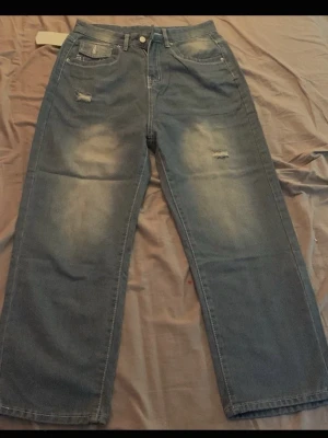 Blå raka jeans från True Religion - Säljer ett par blå jeans från True Religion med raka ben och slitna detaljer på låren. Jeansen har klassiska fickor, vit kontrastsöm och en ikonisk bakficka med lock och broderad logga. Materialet är denim i bomull och passformen är relaxed.