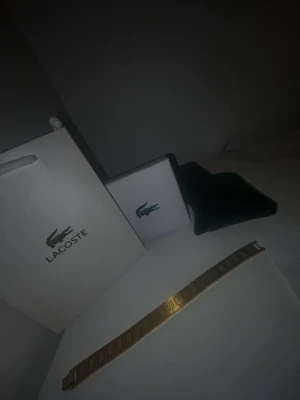 Guldfärgat armband från Lacoste - Snyggt guldfärgat armband från Lacoste med diskret mönster och klassisk design. Armbandet är i metall och har en stilren, rektangulär form med Lacoste-logga. Perfekt accessoar för dig som gillar stilrena detaljer och vill lyfta din outfit.