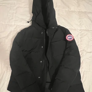 Svart dunjacka från Canada Goose - Säljer en svart dunjacka från Canada Goose med huva och klassisk logotyp på ärmen. Jackan har dragkedja och knappar framtill samt en stilren, puffig design som håller dig varm under kalla dagar. Perfekt för dig som vill ha både stil och funktion.