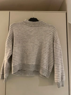 Ljusgrå randig stickad tröja NA-KD - Mysig ljusgrå stickad tröja från NA-KD med tunna horisontella ränder. Tröjan har ribbade muddar vid ärmslut och nederkant samt en högre ribbad krage. Perfekt för lager på lager och chill stil.