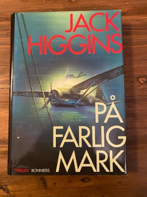 På farlig mark av Jack Higgins - Spännande thrillerbok av Jack Higgins med hård pärm och omslag i blå och gröna toner. Omslaget visar ett flygplan i dramatisk miljö. Boken är utgiven av Bonniers och tillhör genren thriller.