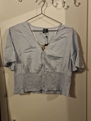 Ljusblå croppad blus från Vero Moda - Söt ljusblå croppad blus från Vero Moda med v-ringning, knappar framtill och korta ärmar. Smockad midja ger en snygg passform och toppen har en luftig och lätt känsla, perfekt för varmare dagar.