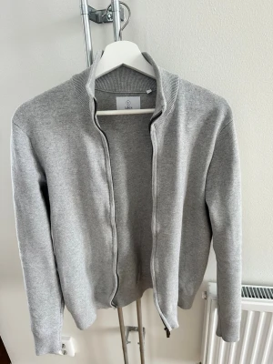 Grå ziptröja från Bläck - Snygg grå ziptröja från Bläck med hög krage och ribbade muddar. Tröjan har dragkedja framtill och är tillverkad i mjuk bomull. Perfekt för lager på lager och enkel att matcha med olika outfits.