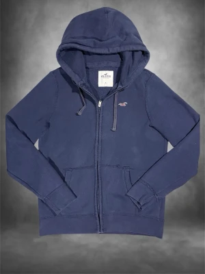 Mörkblå hoodie från Hollister - Mörkblå hoodie med dragkedja från Hollister. Klassisk modell med huva och dragsnören, två fickor framtill och diskret Hollister-logga broderad på bröstet. Tillverkad i mjukt bomullsmaterial som är skönt att ha på sig.