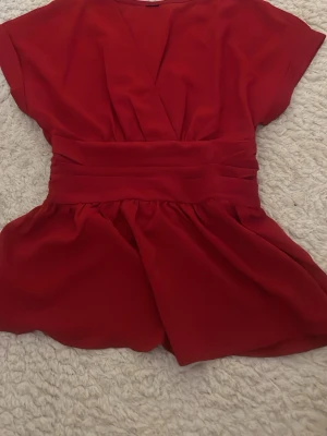 Röd omlottopp med markerad midja - Snygg röd omlottopp med bred markerad midja och korta ärmar. Toppen har ett lätt utsvängt fall nedtill och ett elastiskt parti i ryggen för skön passform. Perfekt för dig som vill ha en feminin och trendig look.