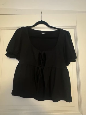 Svart blus med knyt fram - Söt svart blus med puffiga korta ärmar och två knyt framtill. Blusen har en luftig passform och djup rundringning. Perfekt till både jeans och kjol för en trendig look.