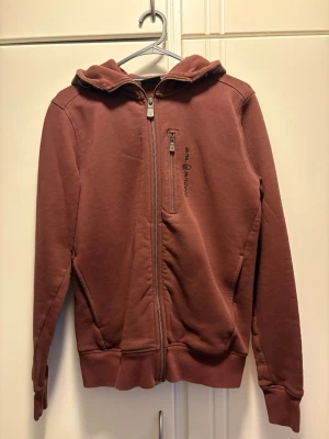 Brun hoodie Sail Racing med dragkedja - Mysig brun hoodie från Sail Racing med dragkedja framtill och en bröstficka med logga. Tröjan har huva, ribbade muddar och två sidofickor. Perfekt för chill dagar och enkel att matcha med jeans eller joggers.