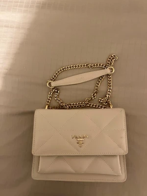 Beige quiltad axelväska - Helt ny! Snygg beige axelväska. Väskan har en stilren, rektangulär form och lockstängning. Perfekt för dig som vill ha en lyxig och trendig accessoar.