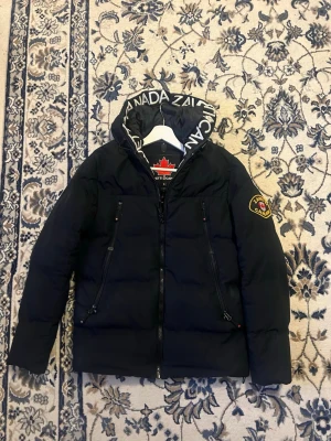 Svart pufferjacka från Zavetti Canada - Säljer en svart pufferjacka från Zavetti Canada med stor huva och vit logotyptext. Jackan har två dragkedjeförsedda fickor framtill och patch på ärmen. Perfekt för kalla vinterdagar och riktigt snygg streetstil.