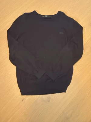 Svart tröja från Hugo Boss - Snygg svart tröja från Hugo Boss med diskret broderad logga på bröstet. Tröjan har rund halsringning, långa ärmar och ribbade muddar vid ärmslut och nederkant. Perfekt för en stilren och enkel look.