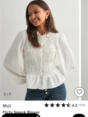 Vit smockad blus från Nelly - Trendig vit blus med smockad front och volang nertill. Blusen har långärmade puffärmar och knappar framtill. Tillverkad i ett lätt och luftigt tyg som ger en romantisk vibe. Perfekt att styla med jeans eller kjol för en chic look.