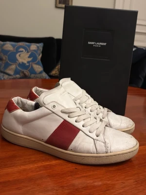 Saint Laurent SL/02 vita och röda sneakers - Snygga vita sneakers från Saint Laurent med röda detaljer på sidan och hälen. De är relativt slitna men har inga vattenläckor och liknande, hälen har dock bra tecken på användning. Va inte rädd att fråga om bilder!