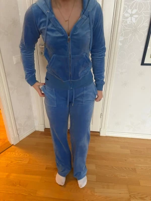 Juicy Couture Blå velour hoodie - Blå hoodie i mjuk velour med dragkedja framtill, huva med snören och två stora fickor. Ribbade muddar vid ärmslut och nederkant ger en skön passform. Perfekt för en chill och trendig look.