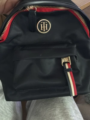 Svart ryggsäck Tommy Hilfiger - Snygg svart ryggsäck från Tommy Hilfiger med guldfärgade detaljer, röd dragkedja och klassiskt TH-emblem framtill. Framtill finns en ficka med dragkedja och dekorativt band i rött, vitt och blått. Tillverkad i slitstarkt material och har en stilren, kompakt form.
