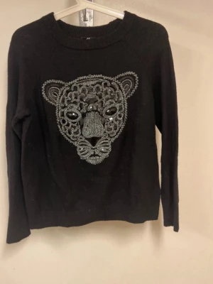 Svart stickad tröja med leopardmotiv - Säljer en svart stickad tröja med ett stort, detaljerat leopardhuvud broderat framtill i grått och silver. Tröjan har rund halsringning, långa ärmar och ribbade muddar. Perfekt för dig som vill ha en cool och unik look.