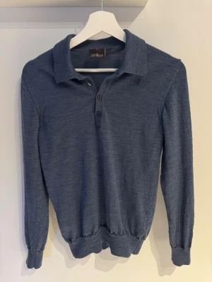 Blå merinotröja från Oscar Jacobson - Snygg blå långärmad tröja från Oscar Jacobson i mjuk merinoull. Tröjan har klassisk krage och tre knappar framtill, samt ribbade muddar vid ärmslut och nederkant. Perfekt för dig som gillar stilrena och bekväma plagg.