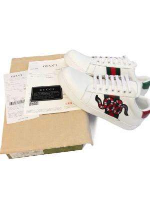 Gucci Ace snake  - Gucci Ace sneakers i vitt läder med klassiska gröna och röda ränder på sidan och en broderad orm i rött, svart och vitt. Hälpartiet har metallicdetaljer i grönt och rött. Snygg och ikonisk modell med vita skosnören och präglad sula.