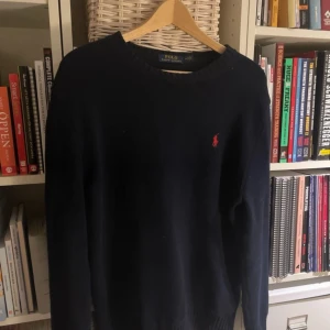 Mörkblå stickad tröja från Polo Ralph Lauren - Klassisk mörkblå stickad tröja från Polo Ralph Lauren med rund halsringning och den ikoniska röda broderade loggan på bröstet. Tröjan har en enkel och stilren design, perfekt för lager-på-lager och snygg till jeans. Använd fåtal gånger, inprincip nyskick. Ny pris ligger runt 1300
