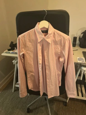 Rosa randig skjorta från Ralph Lauren - Säljer en klassisk rosa skjorta från Ralph Lauren med tunna vita och gröna ränder. Skjortan har lång ärm, knappar framtill och den ikoniska broderade loggan på bröstet. Perfekt för dig som gillar stilrena och tidlösa plagg.