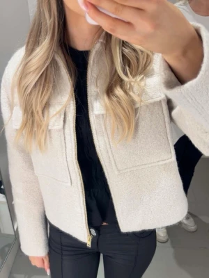 Beige teddyjacka med fickor - Snygg beige med två stora bröstfickor och guldfärgad dragkedja framtill. Jackan har en boxy, croppad passform och är gjord i mjukt, fluffigt material som ger en trendig look. Perfekt för dig som vill ha en snygg jacka till våren! Jackan är från hm, men säljs inte längre. Endast använd vid få tillfällen. 