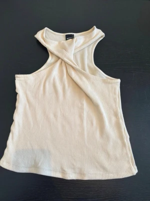 Beige ribbad linnetopp med twistad hals - Trendig beige linnetopp med ribbad struktur och twistad detalj vid halsen. Ärmlös modell med snygg passform, perfekt för varma dagar eller lager på lager. Enkel att matcha med både jeans och kjol.