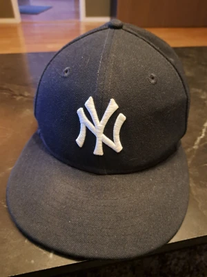 Mörkblå New Era Yankees keps - Snygg mörkblå keps från New Era med klassisk New York Yankees-logga broderad i vitt framtill. Kepsen har rak skärm och ventilationshål på ovansidan. Materialet är tåligt tyg som ger en sportig och stilren look. Perfekt för dig som gillar streetwear och baseball.