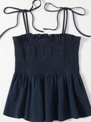 Mörkblå linnetopp med knytband - Söt mörkblå linnetopp med smockad överdel och tunna axelband som knyts upptill. Toppen har en lätt utsvängd nederdel och volangkant upptill, perfekt för en avslappnad och trendig look.