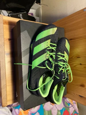 Adidas svarta och limegröna löparspikes - Säljer ett par Adidas löparspikes i svart och limegrönt med stora ränder och coola detaljer. Skorna har metallspikar undertill och en skinande gulgrön sula för max grepp på banan. Perfekta för friidrott och sprint. Sköna och lätta med snörning.