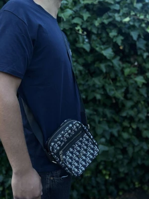  axelväska med monogram - Säljer en snygg axelväska från  med klassiskt svart och grått monogrammönster. Väskan har två dragkedjefack framtill och justerbar axelrem i svart textil. Perfekt storlek för det viktigaste och ger en trendig vibe till din outfit.