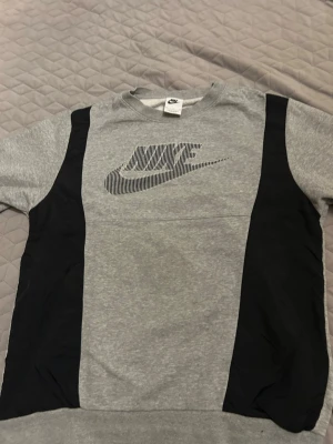 Grå och svart Nike sweatshirt - Snygg grå sweatshirt från Nike med svarta paneler på sidorna och ett stort Nike-logotyptryck i svart på bröstet. Tröjan har rund halsringning och långa ärmar. Perfekt för en sportig och avslappnad stil.