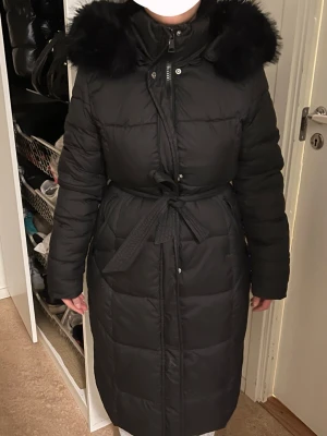 Svart lång dunjacka med fuskpäls - Snygg svart lång dunjacka med bälte i midjan och stor luva med fluffig svart fuskpäls. Jackan har dragkedja och knappar framtill samt quiltad design. Perfekt för kalla dagar när du vill hålla dig varm och stilren.Möts hällre upp, vi finns i stockholm/södertälje.