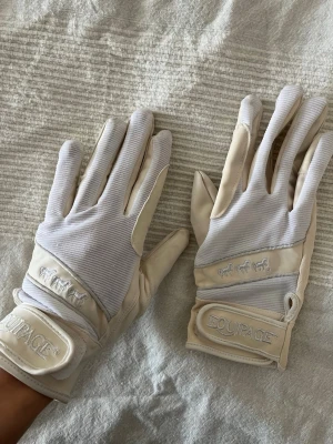 Vita ridhandskar från Equipage - Snygga vita ridvantar från Equipage med broderad logga och hästmotiv på ovansidan. Vantarna har justerbar kardborrestängning vid handleden och är tillverkade i ett flexibelt material med förstärkta partier i handflatan. Perfekta för tävling.