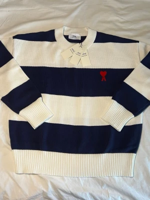 Randig tröja från Ami Paris - Säljer en tröja från Ami Paris med breda marinblå och vita ränder. Tröjan har rund hals, ribbade muddar och nederkant samt Ami-loggan med rött hjärta broderad på bröstet. Perfekt för dig som gillar klassisk men trendig stil.