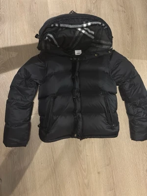 Svart buberry pufferjacka  - Snygg svart pufferjacka med stor vaddering och dragkedjor på fickorna. Jackan har en avtagbar huva med grått och vitt randigt innerfoder. Perfekt för kalla dagar och riktigt trendig look.