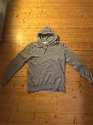 Grå cashmere hoodie från Sayless - Mjuk och lyxig grå hoodie från Sayless i 100% cashmere. Klassisk modell med huva och dragsko, ribbade muddar och avslappnad passform. Perfekt för dig som gillar stilrena och bekväma plagg.