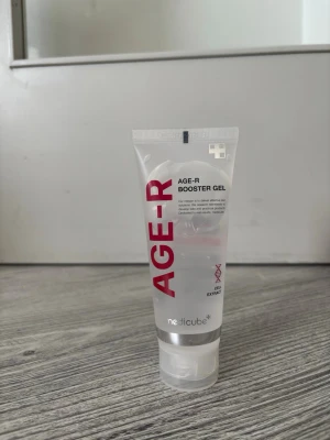AGE-R Booster Gel från Medicube - AGE-R Booster Gel från Medicube är en genomskinlig gel i en plasttub med röd och svart text. Gelen är framtagen för att boosta hudens återfuktning och innehåller kollagenextrakt. Tuben har en praktisk flip-top-kork och rymmer en generös mängd produkt.