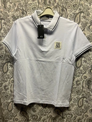 Vit pikétopp från Armani Exchange - Vit pikétopp från Armani Exchange med svart-vita ränder på krage och ärmslut. Klassisk polokrage, kort ärm och broderad logotyp på bröstet. Tillverkad i bomull för en fräsch och sportig look.